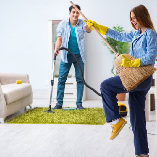 A woman holds a broom, pretending to play it like a guitar, standing on one leg. She wears yellow cleaning gloves. In the background, a man vacuums the green rug, adding a touch of professional carpet cleaning flair. Both are smiling in their bright, tidy living room.