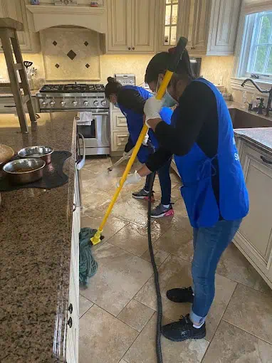 Two people wearing blue aprons are busy with residential cleaning, tackling both the kitchen floor with a mop and vacuum cleaner. The kitchen boasts a large stove, granite countertops, and beige cabinets.