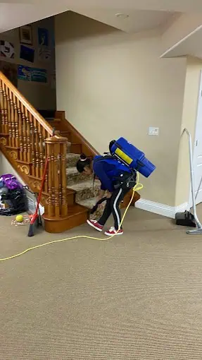 A person with a blue backpack vacuum is diligently performing residential carpet cleaning on a staircase. Nearby, a broom and vacuum cleaner rest on the beige carpeted floor. The stairs, adorned with wooden railings, are in a bright and welcoming room.