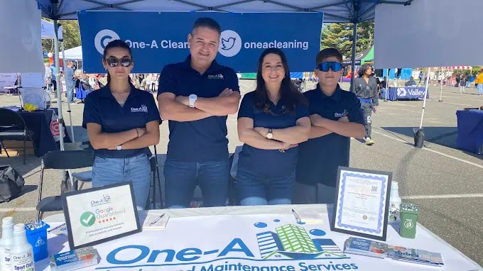 Four people wearing navy shirts with a logo stand confidently behind a booth table at an outdoor event. The booth has a banner that reads "One-A Cleaning Services." Specializing in residential cleaning, the table displays promotional items, business cards, and a framed certificate.