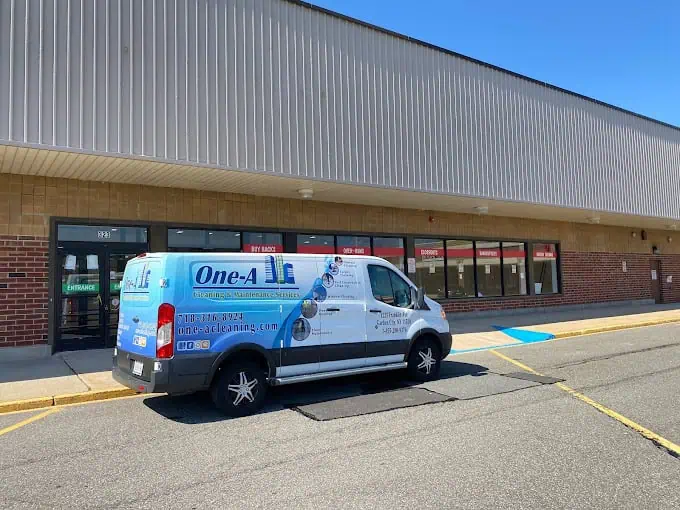 A white van parked in front of a row of closed shops showcases contact information for "One-A Cleaning & Maintenance Services," known for expert residential cleaning. Adorned with images of cleaning tools and a website URL, the vehicle gleams under clear skies in the well-lit area.