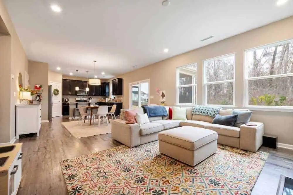 A spacious open-concept living area features a beige sectional sofa with colorful cushions, a patterned rug, and a view of the kitchen with dark cabinets and modern lighting. Large windows let in natural light, highlighting the pristine ambiance maintained by top-notch apartment cleaning services.