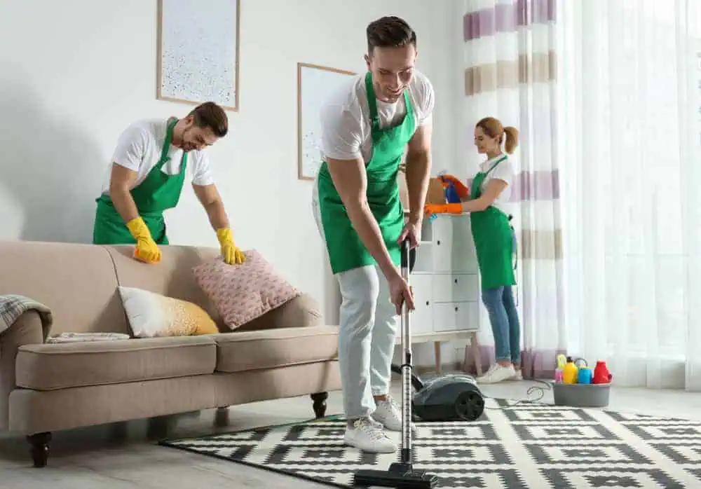 Three people in green aprons are cleaning a living room. One is vacuuming a geometric-patterned rug, another is dusting a pillow on a beige sofa, and the third is cleaning a window. Cleaning supplies are on the floor nearby.