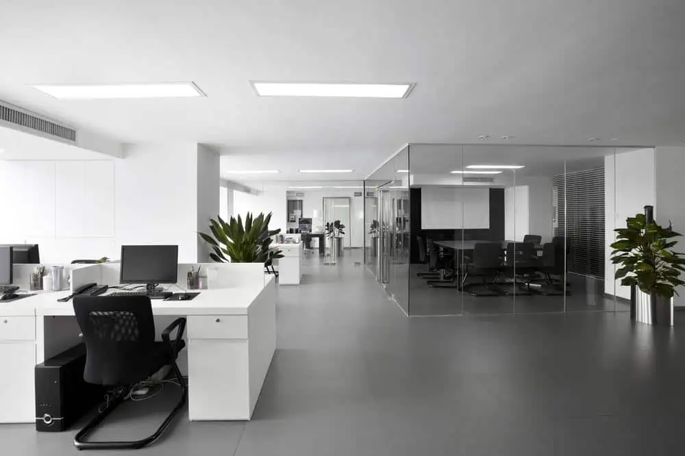 Modern office with white walls and minimalist furniture. The space features open workstations with computers, several indoor plants, and a glass-enclosed meeting room. Bright overhead lighting highlights the sleek design.