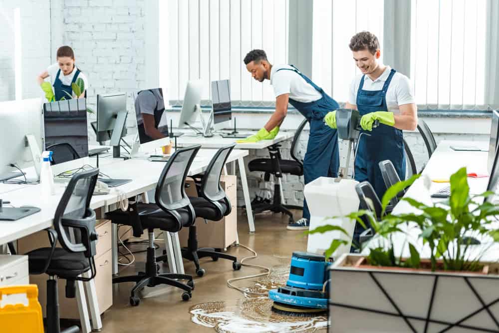 Three people in uniforms are cleaning a modern office. Two are wiping desks and one is operating a floor cleaning machine. The office is bright with large windows, white walls, and several computer workstations. A plant is visible in the foreground.