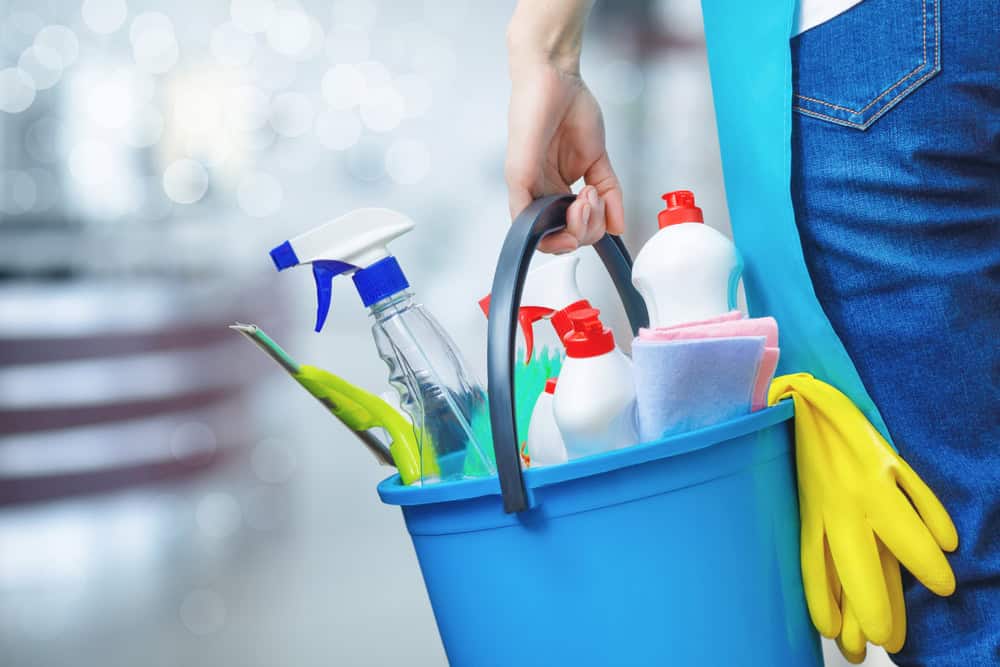 A person holding a blue bucket filled with various cleaning supplies, including spray bottles, sponges, and cloths. Yellow rubber gloves are draped over the side of the bucket. The background is blurred with bright lights.