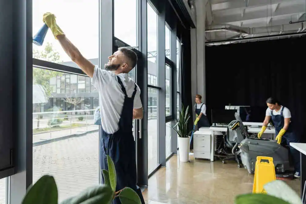 A cleaning crew works in a modern office with large windows. One person cleans a window, while another tidies a desk, and a third vacuums. A caution wet floor sign is visible. The room has plants and sleek furniture.