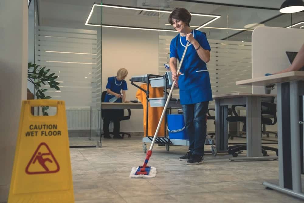 A person in a blue uniform mops an office floor, with a "Caution: Wet Floor" sign nearby. Another person pushes a cleaning cart in the background. The office has modern glass partitions and desks.