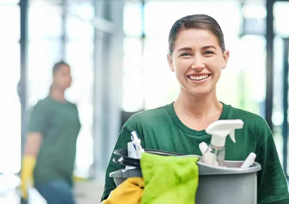 A woman in a green shirt smiles while holding a cleaning caddy filled with supplies. In the blurred background, a person in a similar outfit is cleaning with yellow gloves. Bright and airy environment.