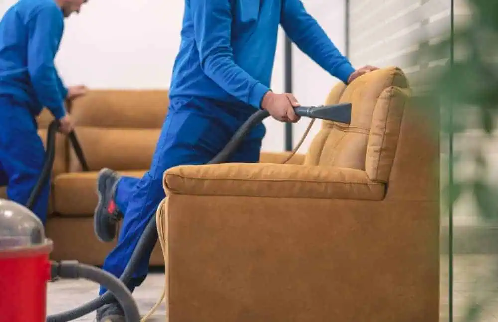 Two people in blue uniforms are cleaning a brown sofa with vacuum cleaners. They focus on different parts of the sofa in a well-lit room. A red vacuum cleaner is visible in the foreground.