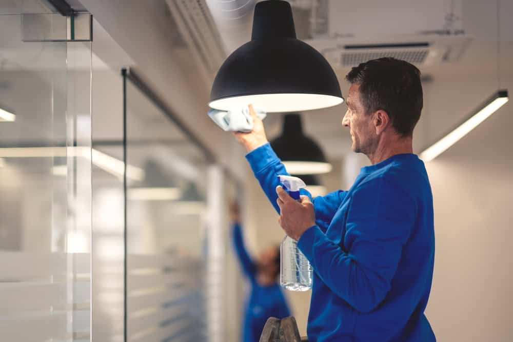 A person in a blue uniform stands on a ladder, cleaning a large black hanging light fixture with a cloth. They hold a spray bottle in the other hand. Another worker is visible in the background, also cleaning. The setting appears to be an office.