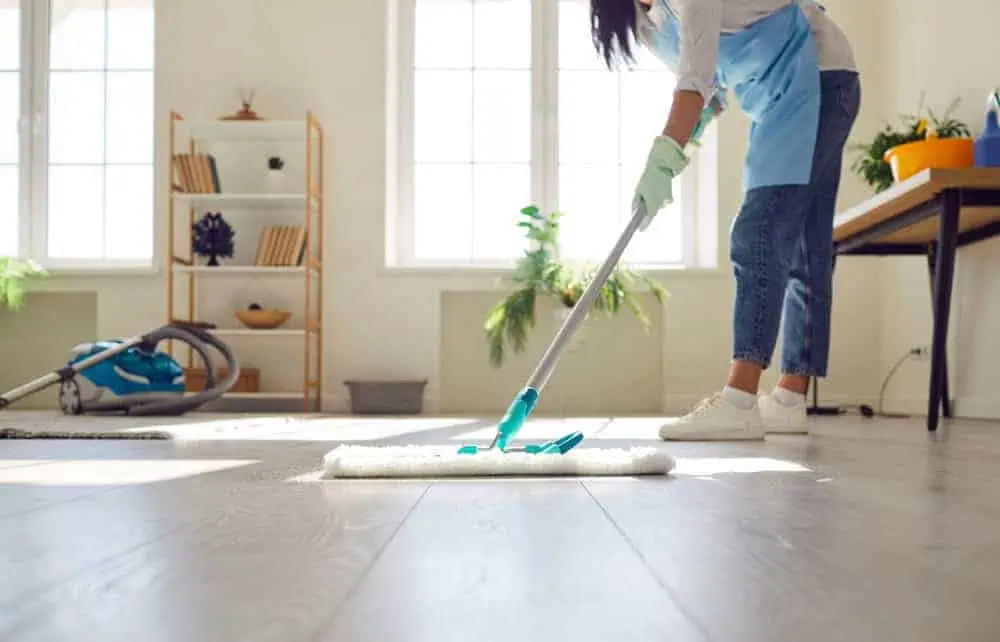 A person wearing a blue apron and jeans is mopping a bright, sunlit room with wooden floors. A vacuum cleaner and shelf with books and plants are visible in the background.