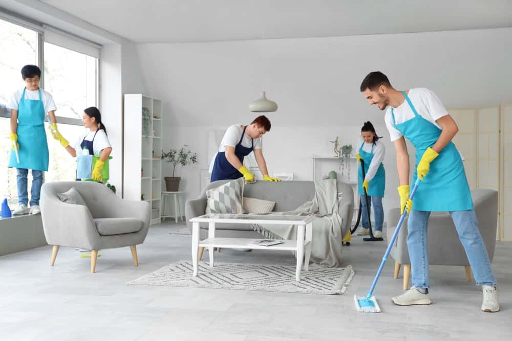 A group of five people in blue aprons and yellow gloves are cleaning a bright, modern living room. Some are wiping surfaces, while others mop the floor. The room has gray furniture, a white coffee table, and large windows letting in natural light.
