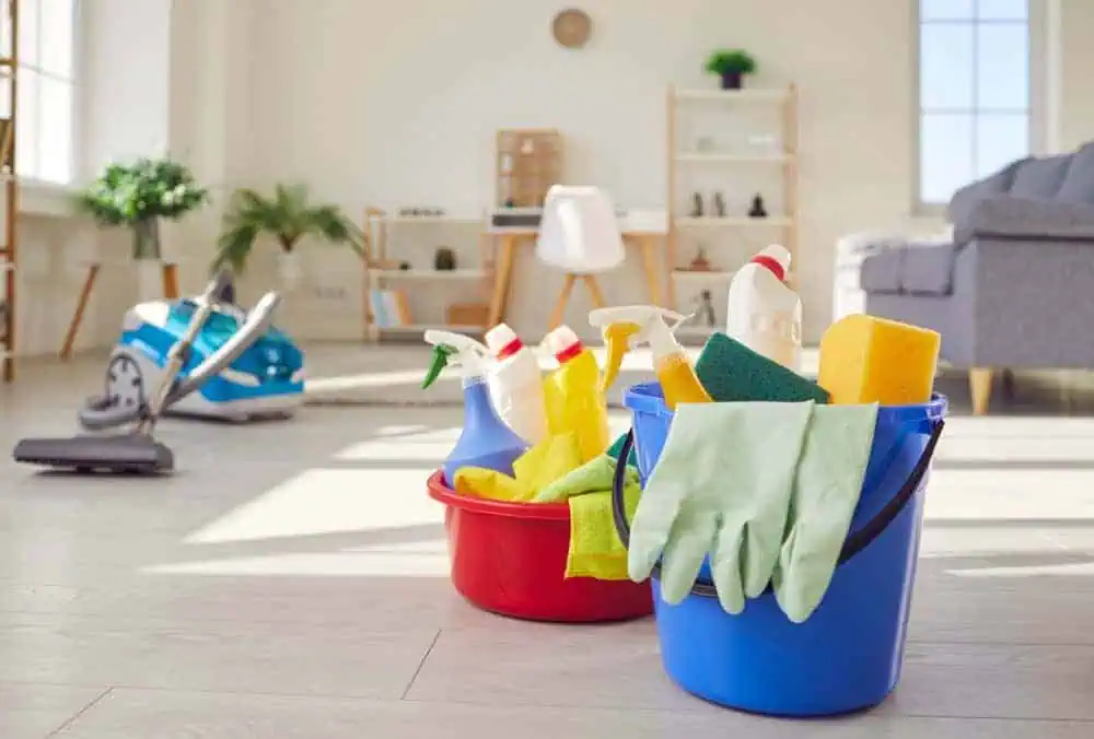 A clean, sunny living room with two colorful buckets containing cleaning supplies like spray bottles, sponges, and rubber gloves. A vacuum cleaner is in the background, next to a plush gray sofa and a minimalist desk with a chair.