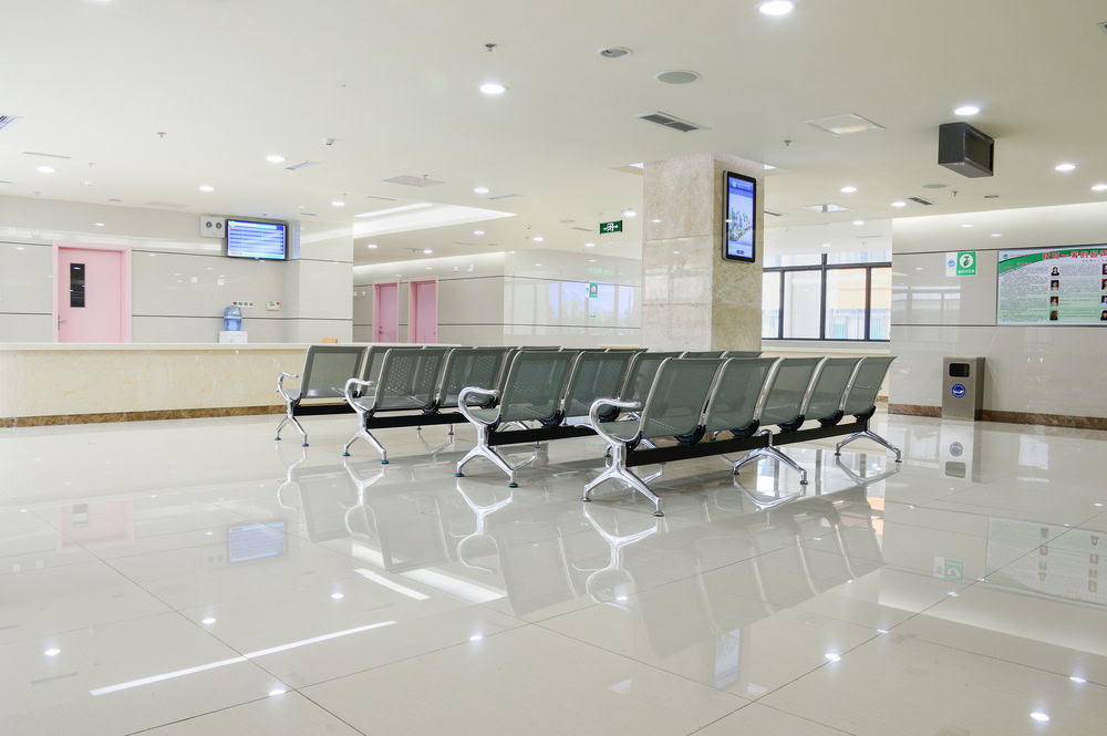 A modern, empty waiting area in a hospital or clinic. Rows of metal chairs are aligned neatly on a reflective tiled floor. Digital screens and informational posters are visible on the walls, with pink doors in the background.