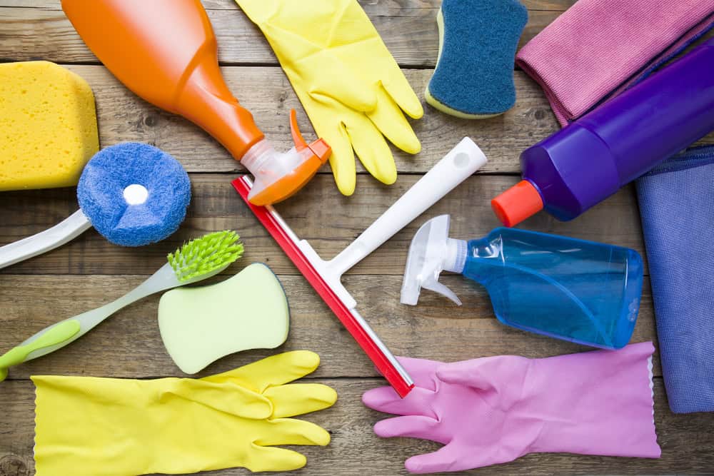 A collection of cleaning supplies on a wooden surface, including a squeegee, spray bottles, sponges, a scrub brush, and rubber gloves in yellow and pink. Various bottles and scrub pads in orange, blue, and purple are also visible.
