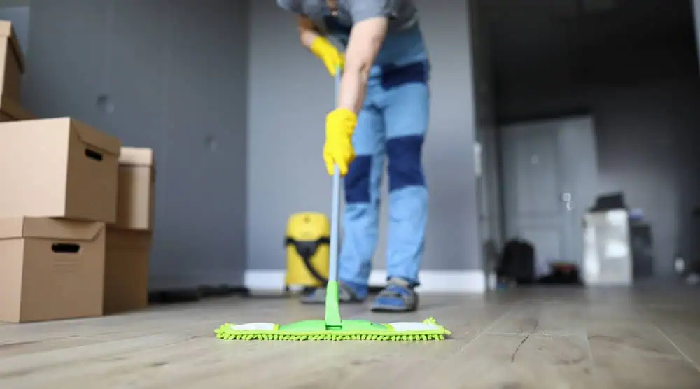 A person wearing yellow gloves is mopping a wooden floor in a room with cardboard boxes. The person is dressed in blue and appears to be cleaning or organizing a space, possibly during a move or renovation.