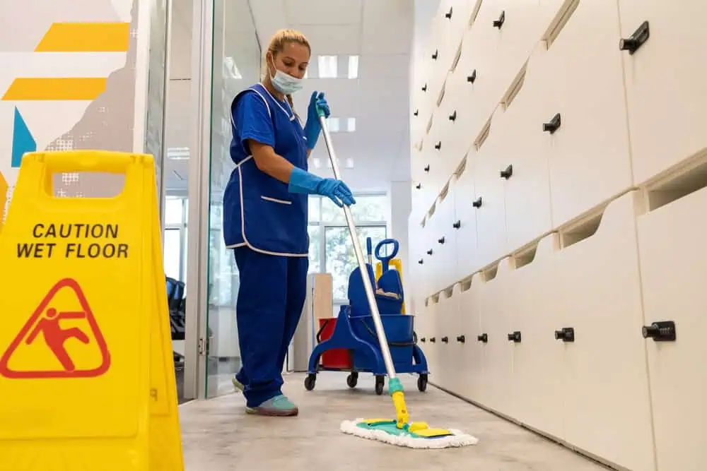 A cleaning professional mops a floor in an office hallway, wearing a blue uniform, gloves, and a face mask. A "Caution Wet Floor" sign is visible on the left. Cleaning supplies are in the background. White lockers line the right side.