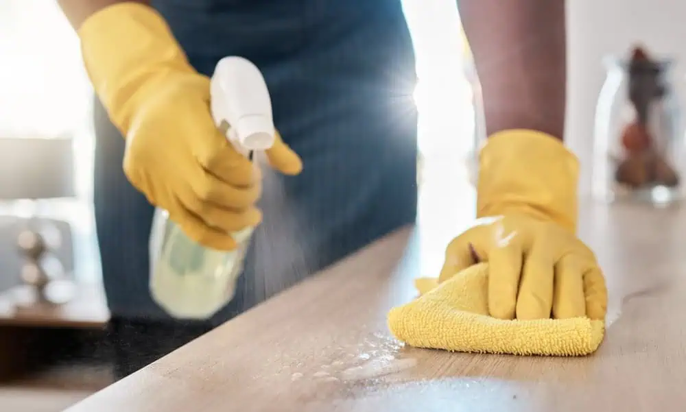 A person wearing yellow rubber gloves is cleaning a surface with a spray bottle and a yellow cloth. Sunlight shines through a window, illuminating the scene.