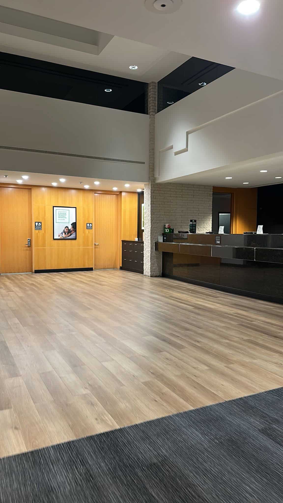 Modern lobby with wooden flooring, a dark reception desk on the right, and wood-paneled doors straight ahead. The space features a white ceiling with recessed lighting and a framed photo on the wall near the doors.