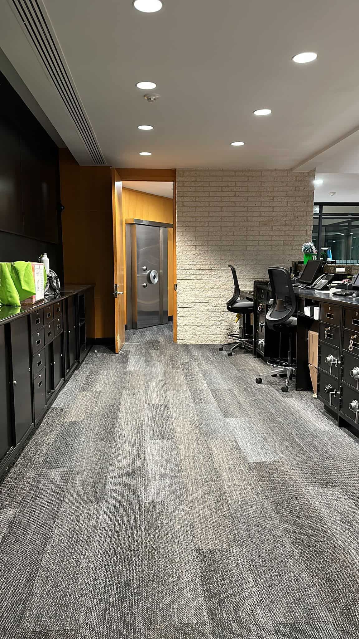 Office space with gray carpet, black cabinets, and swivel chairs. A wooden door is open, revealing a vault with a large metal door. Bright overhead lights illuminate the room. A green bag is on a cabinet.