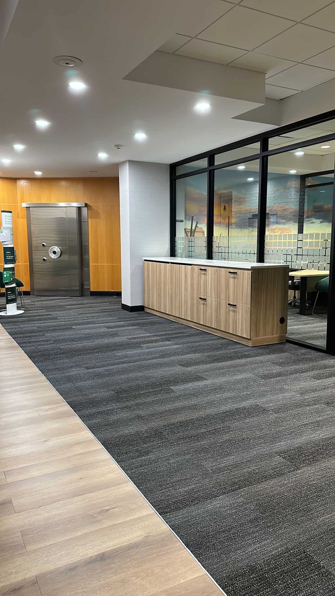 A modern office lobby featuring a wooden cabinet on a gray carpeted floor. The room has bright overhead lights, a large vault door on the left, and a window showing a scenic sunset mural.