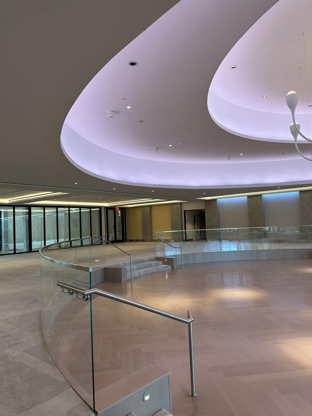 Spacious and modern interior with a large, curved ceiling featuring recessed lighting. A glass railing follows the curve of the hallway. The room is well-lit and has a minimalist design with light-colored flooring and walls.