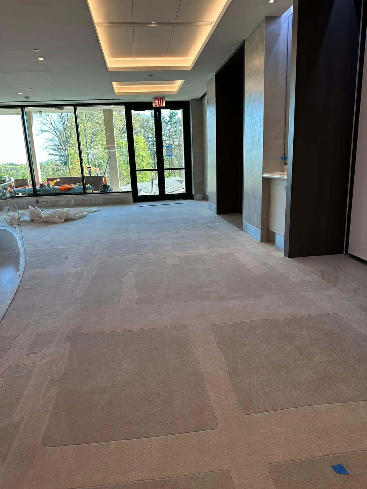A modern corridor with light gray carpet and white ceiling panels. Glass doors at the end lead to an outdoor area with trees visible. Natural light streams in, illuminating the space. A small lounge area with cushions is to the left.