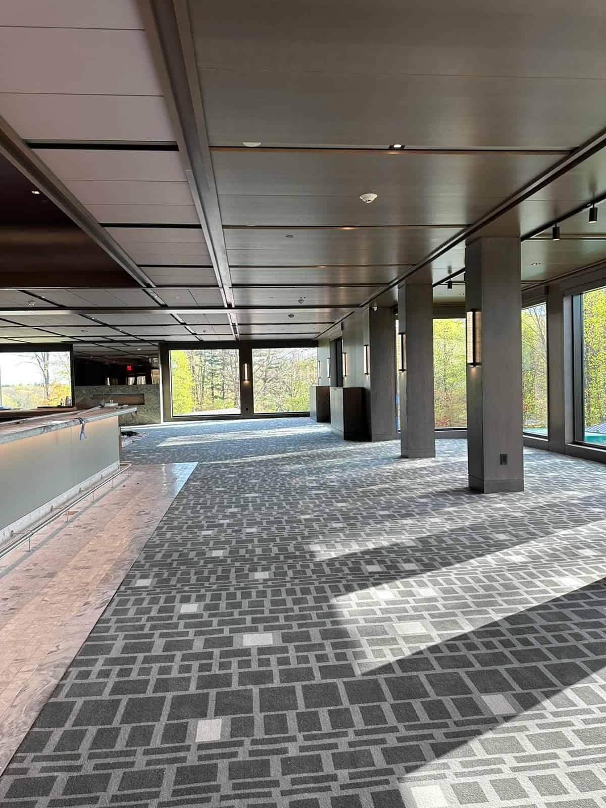 Spacious, modern interior with a geometric-patterned carpet, large windows providing natural light, and a ceiling with recessed lighting. A long counter is on the left, and trees are visible outside the windows.