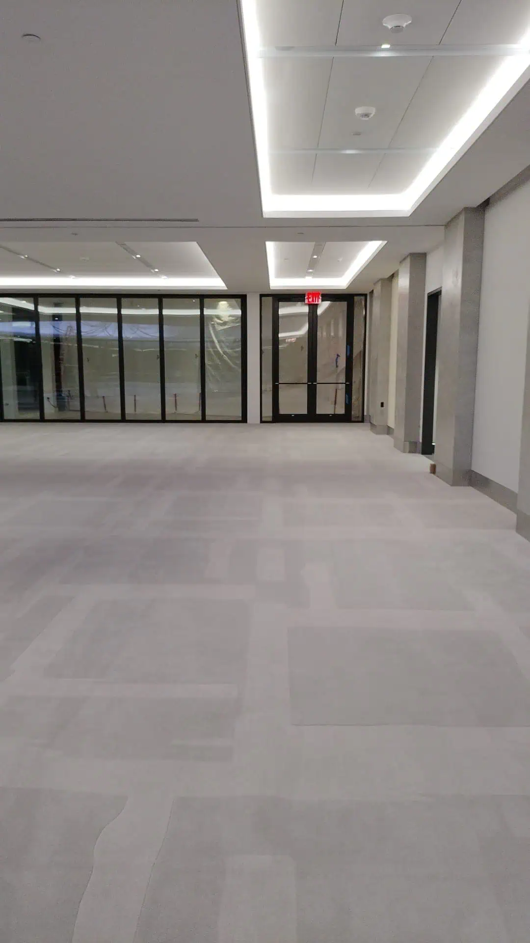 A spacious, empty room with gray carpet and a modern ceiling with recessed lighting. Large glass doors line one wall, revealing a corridor. The room feels clean and contemporary, with minimal decor.