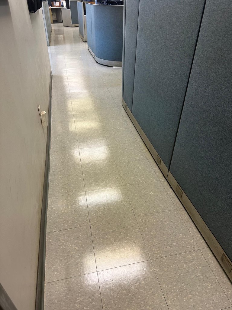 A hallway in an office with light gray tiled floors, blue-gray cubicle walls on the right, and a plain wall on the left. The area is well-lit with fluorescent lights reflecting on the floor.