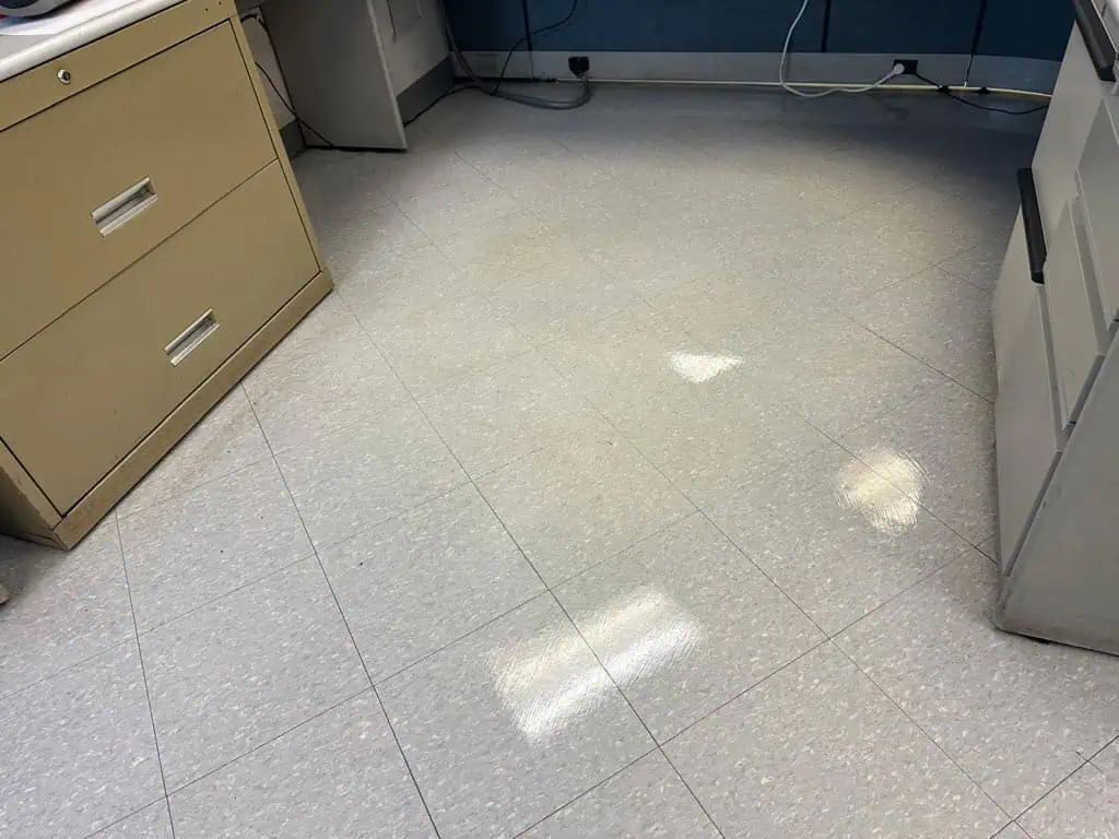 A clean, shiny office floor with light gray tiles, reflecting overhead lights. Filing cabinets, a desk, and cords are visible along the edges of the room.