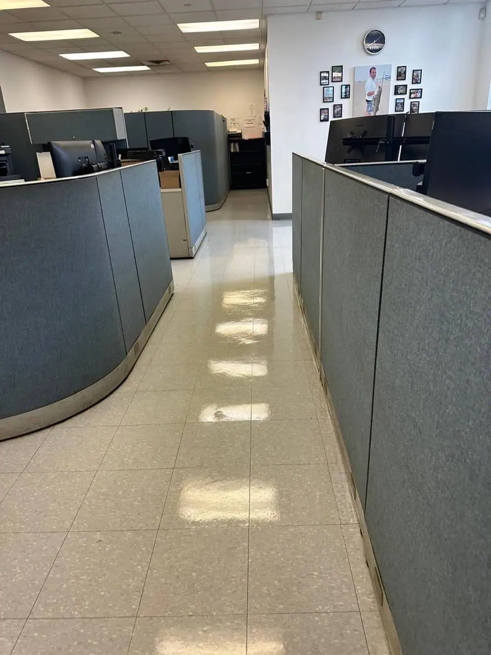 A clean, empty office space with gray cubicles, tiled floor, several computer monitors, and framed photos on a white wall in the background. Fluorescent lights are reflected on the floor.