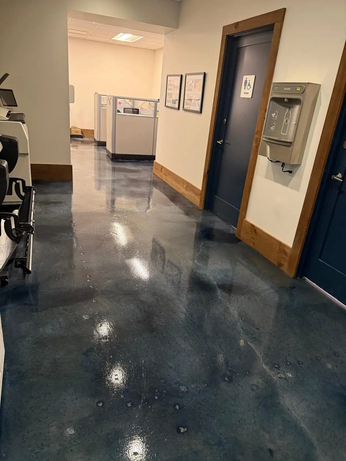 A hallway with shiny dark blue floors, doors on either side, cubicles visible at the end, framed pictures on the wall, and a wall-mounted telephone near a restroom sign.