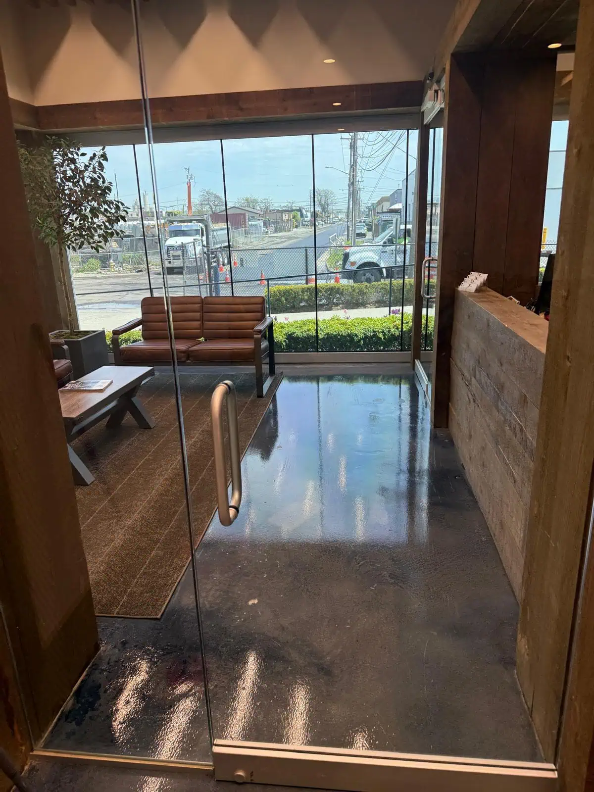 A modern office lobby with a glass door, wooden reception desk, leather sofa, coffee table, and large windows showing an outdoor view with vehicles, power lines, and a sunny sky. The polished floor reflects the light.