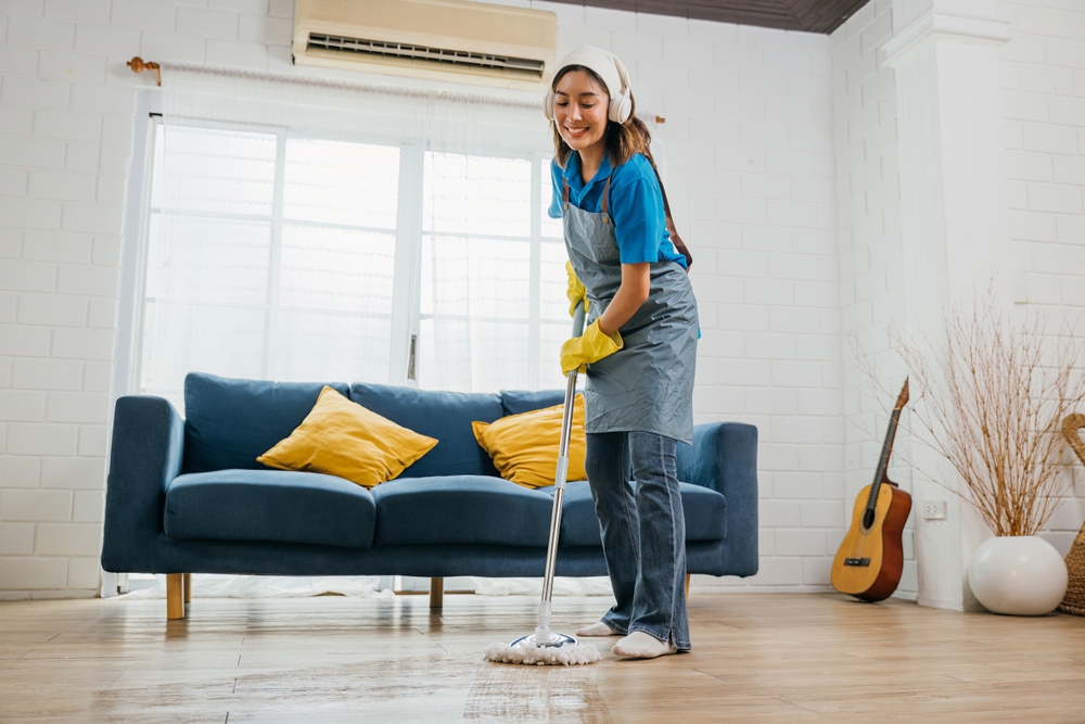 A woman in jeans, a blue shirt and a grey apron mopping a hard wood floor in a living room. She is wearing white headphones and a white bandana. Behind her there is a blue couch with yellow pillows. In the corner, there is an acoustic guitar leaning against the white wall. On the other side of the corner, there is a circular, white pot with branches coming out.