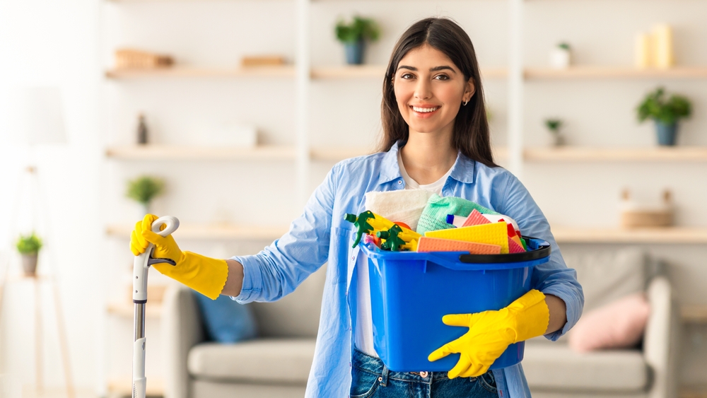 A woman in a living room wearing jeans, a white shirt and a light blue button down. Her sleeves are rolled up to her forearms and she is wearing yellow rubber gloves. One hand holds a blue bucket full of cleaning supplies and the other is holding onto the handle of a mop.