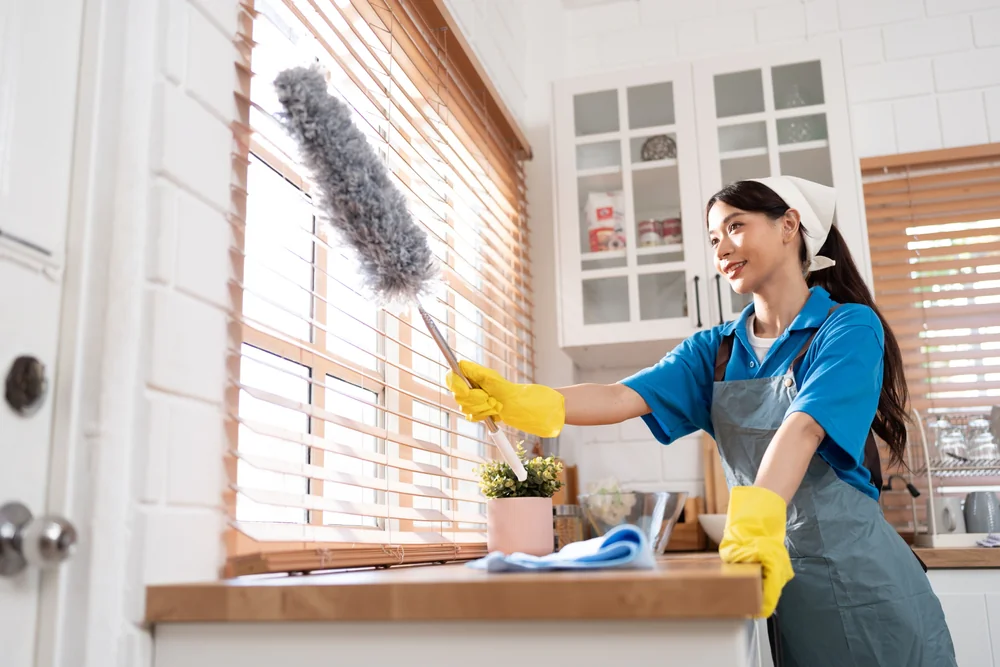 A woman in NY wearing a blue shirt, apron, and yellow gloves dusts wooden window blinds in a bright kitchen. Cleaning services Nassau County supplies and a potted plant sit on the countertop beside her.