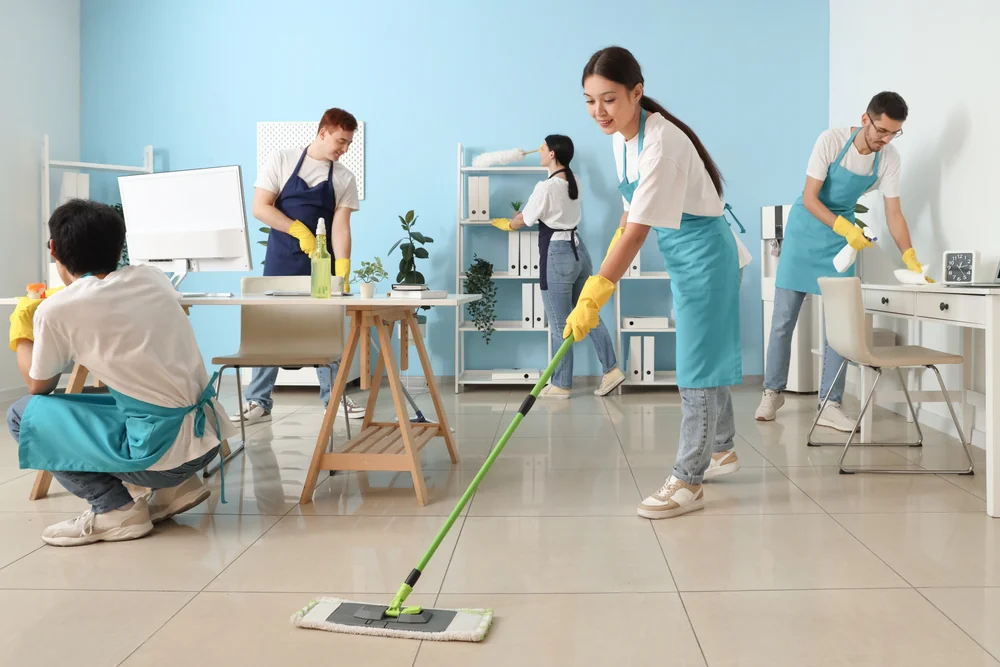 A team of four people wearing aprons and yellow gloves clean a modern NY office, wiping surfaces, mopping the floor, and organizing shelves. They provide top-notch cleaning services Nassau County relies on for spotless workplaces.