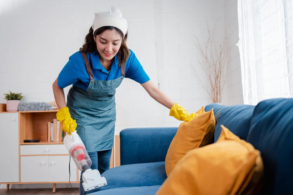 A woman wearing a blue shirt, apron, and yellow gloves is smiling while vacuuming a blue sofa with yellow cushions in a bright living room. Sunlight streams through a window—showcasing top cleaning services Nassau County, NY.