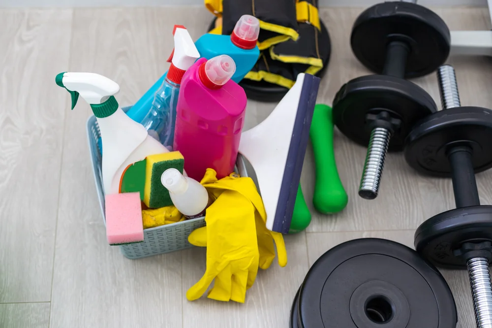 A basket of cleaning supplies, including spray bottles, sponges, rubber gloves, and a squeegee, sits on a light-colored floor next to several black dumbbells and weight plates.
